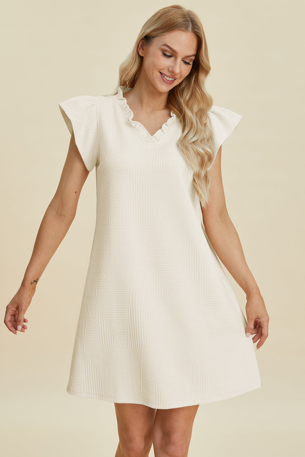 A woman smiles while looking down in the cream-colored Double Take Full Size Ruffled V-Neck Cap Sleeve Dress made of polyester. Her long blonde hair cascades over her shoulders against a plain beige background. This easy-care dress is perfect for everyday wear with machine wash cold instructions.