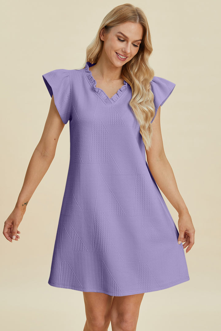 A person in a lavender Double Take Full Size Ruffled V-Neck Cap Sleeve Dress featuring a textured geometric pattern, crafted from easy-care, machine-washable cold polyester, stands against a light beige backdrop, smiling with arms slightly outstretched.