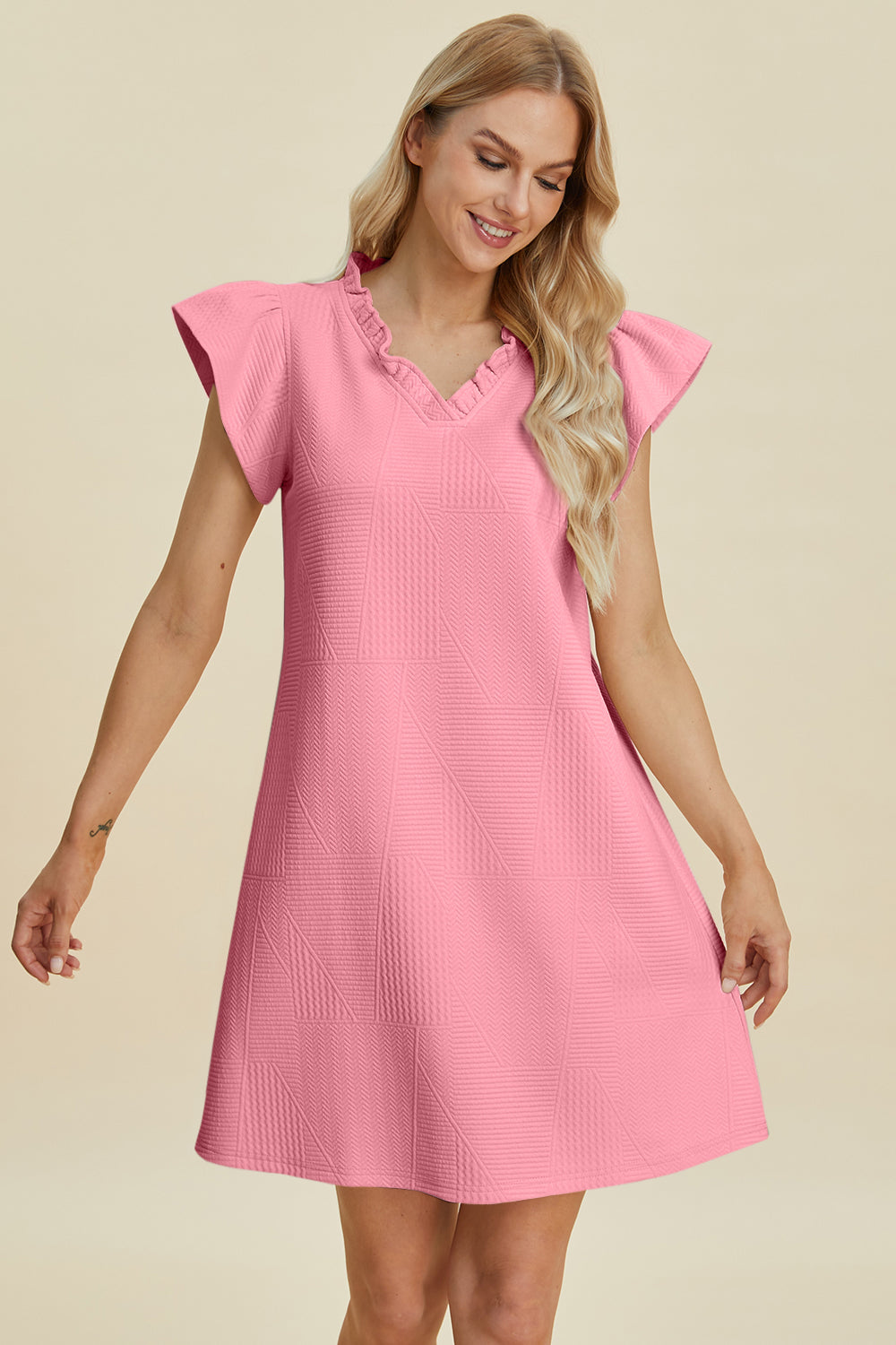 A woman wearing the Double Take Full Size Ruffled V-Neck Cap Sleeve Dress, made of pink textured polyester with a ruffled neckline and cap sleeves, smiles downwards against a beige background. Elegantly practical for daily wear; remember to machine wash cold to maintain freshness.