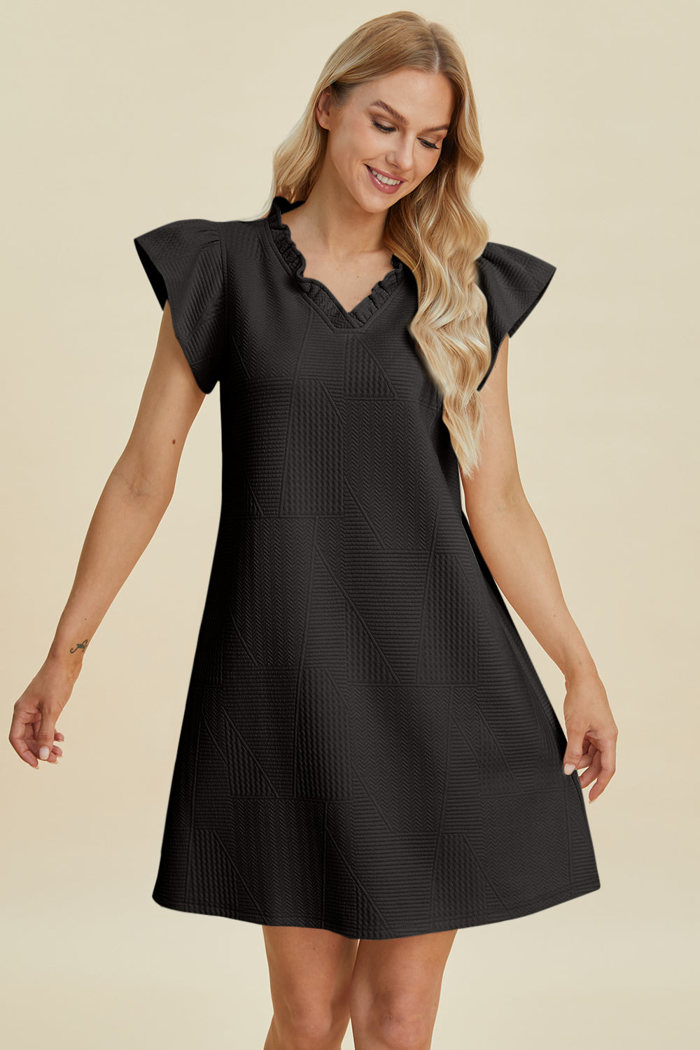 A woman with long blonde hair wears the Double Take Full Size Ruffled V-Neck Cap Sleeve Dress in black polyester, standing against a plain background, smiling and looking down.