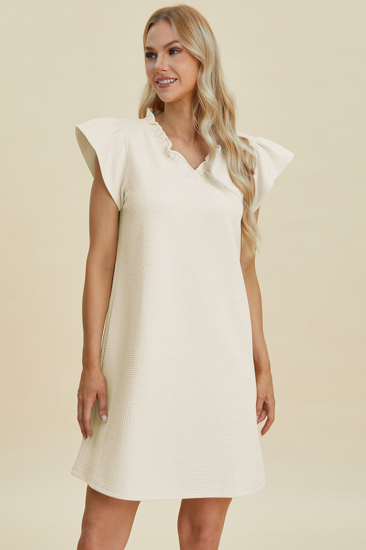 Smiling, a person wears the Double Take Full Size Ruffled V-Neck Cap Sleeve Dress in cream polyester with short, ruffled sleeves. The loose fit drapes above the knees, and their long, wavy hair complements the soft cream background. Machine wash cold for care.