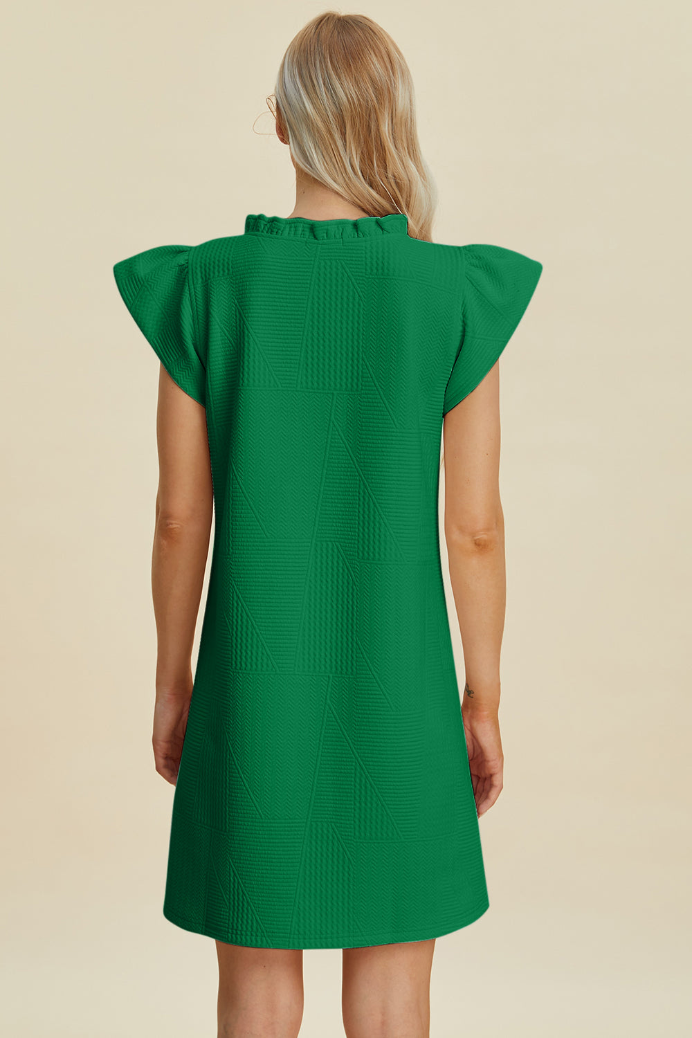 A person with long blonde hair is seen from behind wearing the Double Take Full Size Ruffled V-Neck Cap Sleeve Dress in green. Made of polyester, it features ruffled edges on the shoulders and collar with a textured pattern in a basic style against a plain beige background.