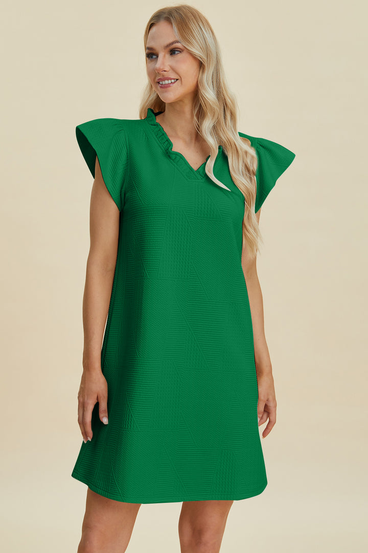 A person wearing the Double Take Full Size Ruffled V-Neck Cap Sleeve Dress in green polyester, featuring a subtle geometric pattern, stands against a beige background. They have long, wavy blonde hair and are smiling to the side. Easy-care with machine wash cold instructions, ideal for basic style lovers.