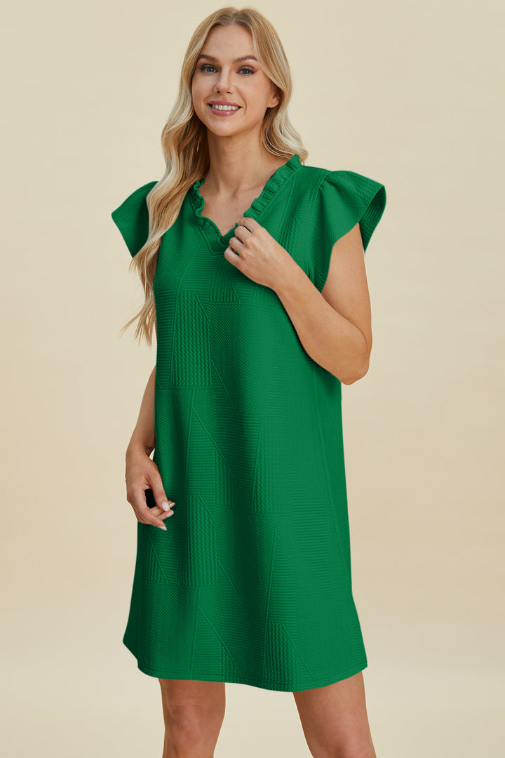 A person with long blonde hair smiles, wearing the Double Take Full Size Ruffled V-Neck Cap Sleeve Dress in green. This polyester dress is knee-length with a ruffled neckline and short sleeves, offering easy care with machine wash cold. They stand against a plain beige background.