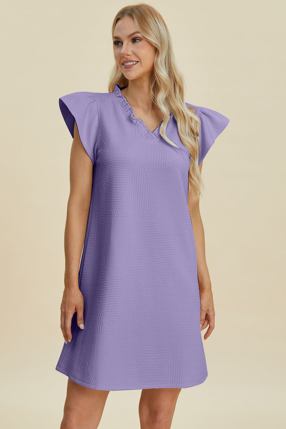 A person smiles against a plain, light background in a Double Take Full Size Ruffled V-Neck Cap Sleeve Dress. Made of textured purple polyester, it has cap sleeves and is machine washable. They have long blonde hair and stand with one hand by their side.