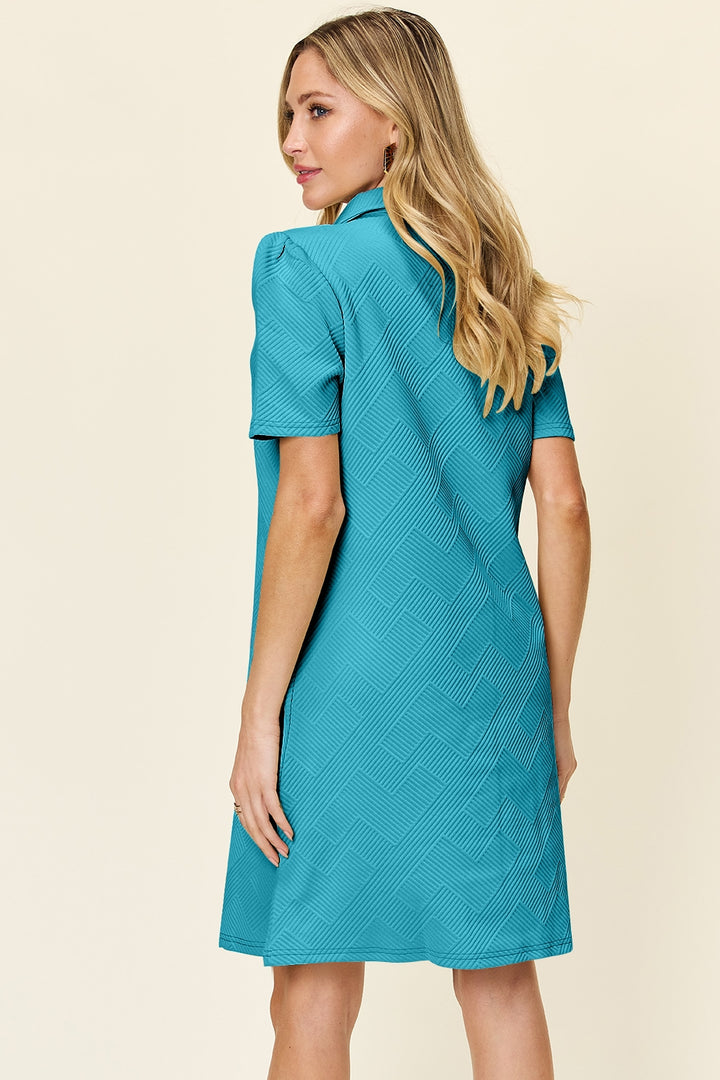 A woman with long blonde hair stands in a Double Take Full Size Texture Collared Neck Short Sleeve Dress, made from imported teal polyester and featuring a subtle geometric pattern. She is smiling and facing slightly to the side against a plain light background.