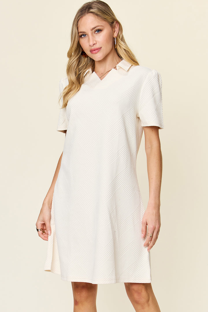 A woman in the Double Take Full Size Texture Collared Neck Short Sleeve Dress, featuring a cream-colored design with a subtle checkered pattern that highlights her basic style. Her long, wavy blonde hair cascades gracefully as she stands against a light beige background, looking confidently at the camera. The dress is crafted from imported polyester for a polished finish.