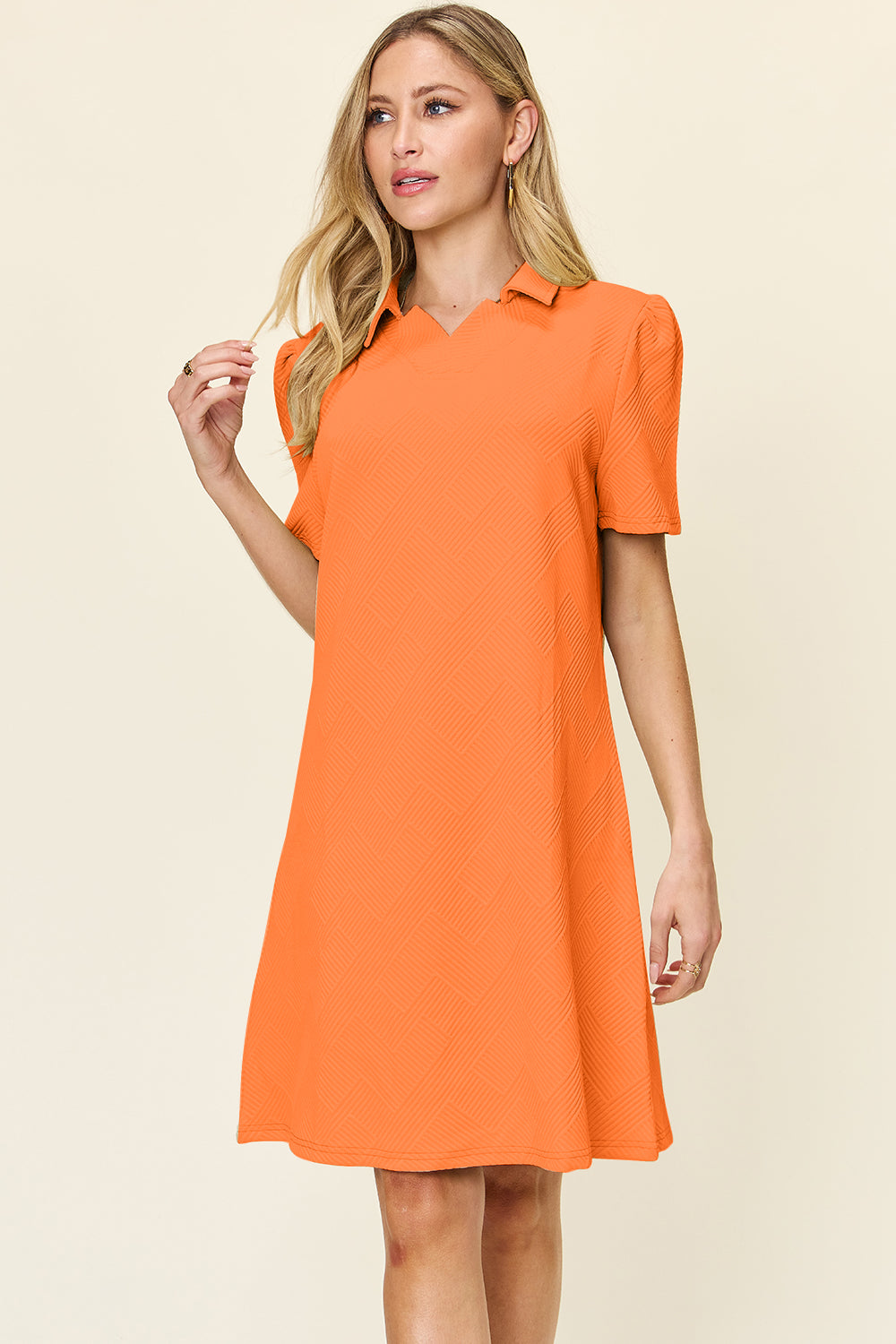 A woman wearing the Double Take Full Size Texture Collared Neck Short Sleeve Dress, a vibrant orange polyester dress featuring short sleeves and a collar, stands against a plain background. Her long, blonde hair cascades down as she gazes thoughtfully to the side, embodying a basic style. With her left hand gently touching her imported locks, she appears lost in thought.