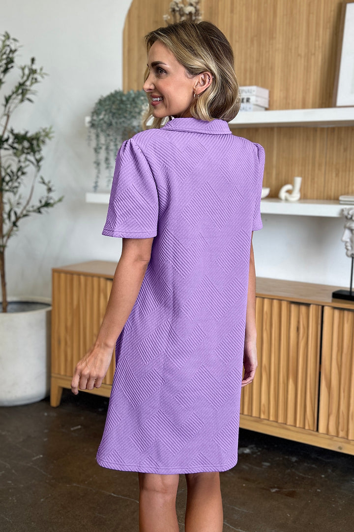 Wearing a Double Take Full Size Texture Collared Neck Short Sleeve Dress in purple, made from imported polyester, a person stands amidst wooden cabinets, white shelves, and potted plants. They are turned slightly to the side, looking back.