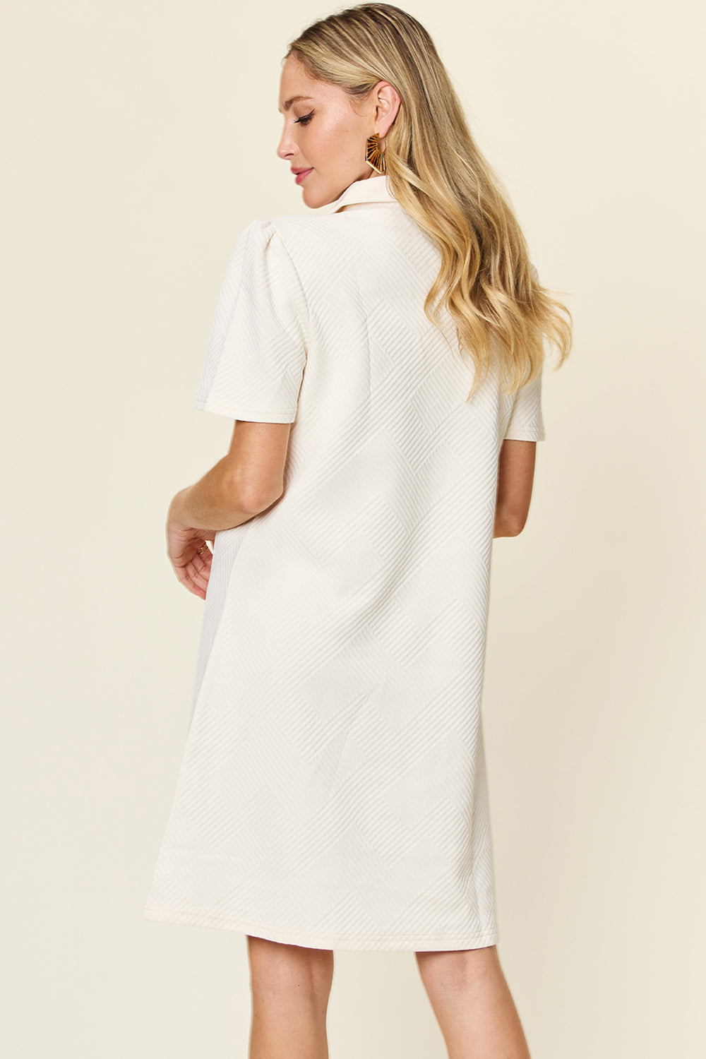 A woman with long blonde hair is wearing the Double Take Full Size Texture Collared Neck Short Sleeve Dress, a knee-length, short-sleeve dress in white made from imported polyester. She is standing and facing away, highlighting the subtle back design pattern. The soft, neutral background complements the basic style of her attire.