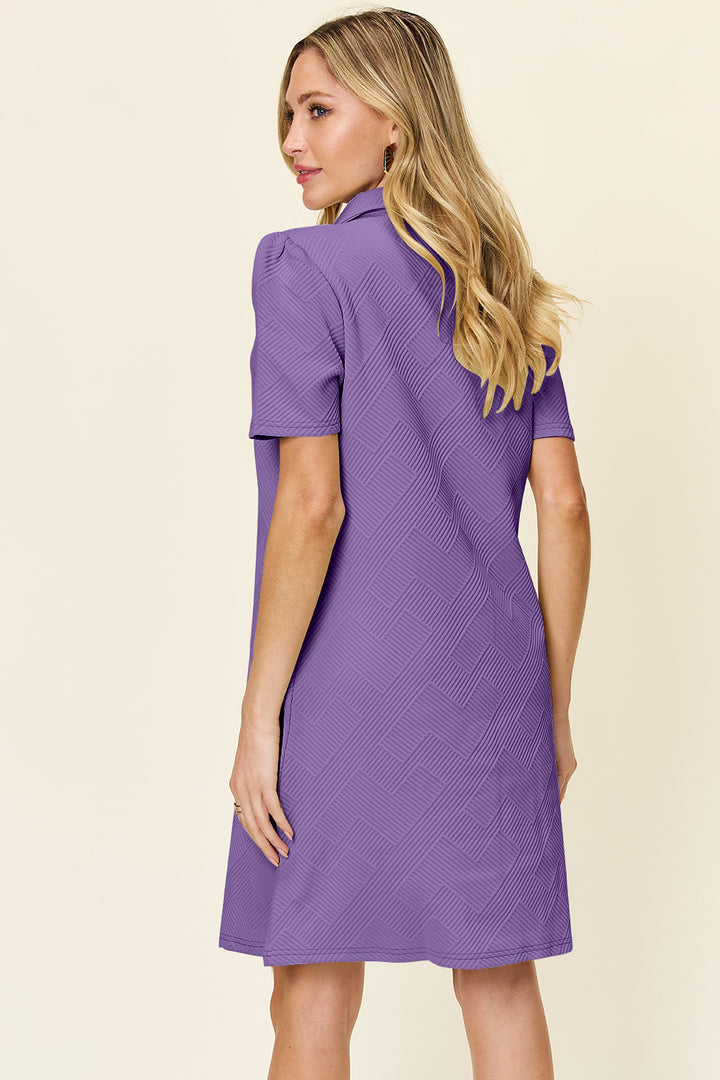 A woman with long blonde hair models the Double Take Full Size Texture Collared Neck Short Sleeve Dress, which is purple and knee-length. Made from imported polyester, this dress features a basic style complemented by a subtle zigzag pattern. She stands with her back to the camera, slightly turning her head to the side.