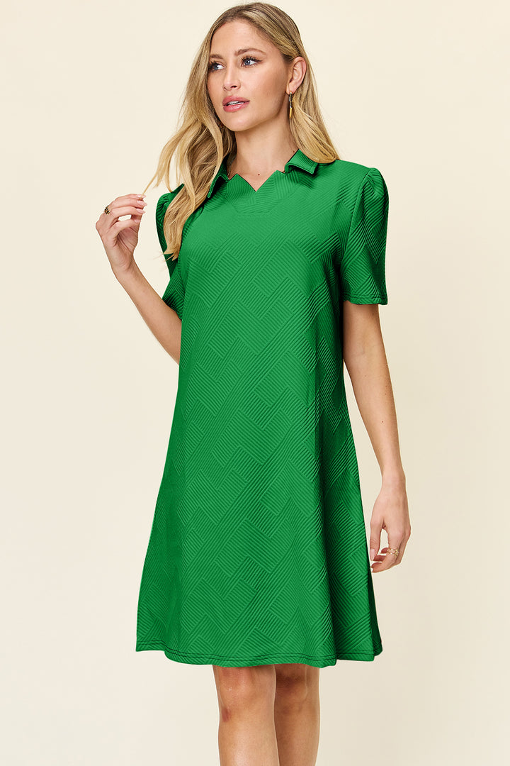 A woman stands against a plain background in the Double Take Full Size Texture Collared Neck Short Sleeve Dress, showcasing its basic style and subtle pattern. Made from imported polyester, the green dress features short sleeves and exudes a relaxed and casual vibe as she gazes to the side with her hand near her hair.