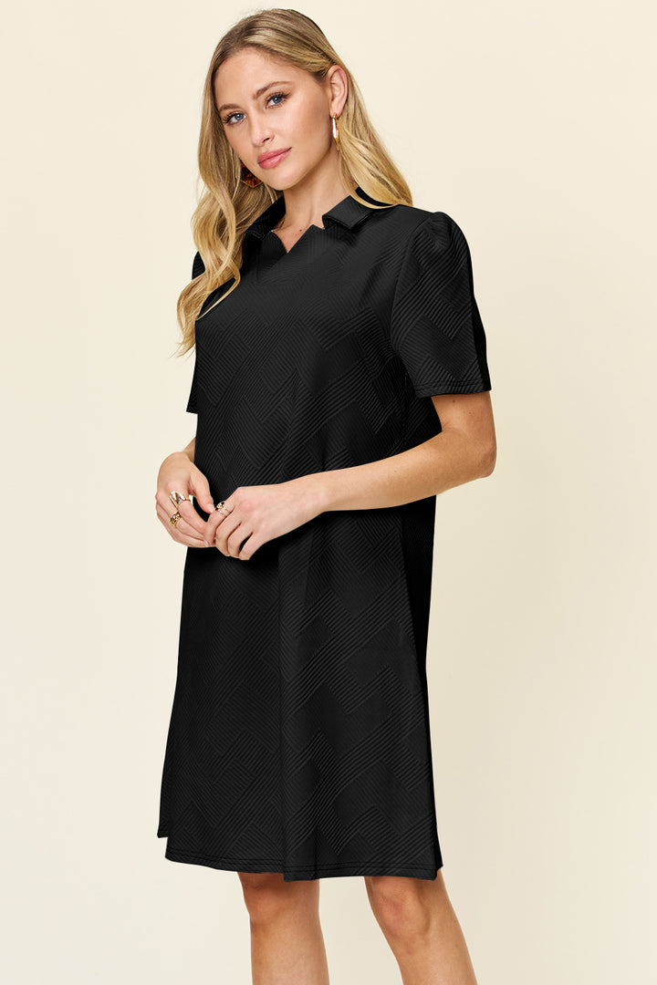 A woman with long hair is wearing the Double Take Full Size Texture Collared Neck Short Sleeve Dress, made of imported polyester in black, and holds her hands together. The plain light-colored background accentuates the basic style of her elegant attire.