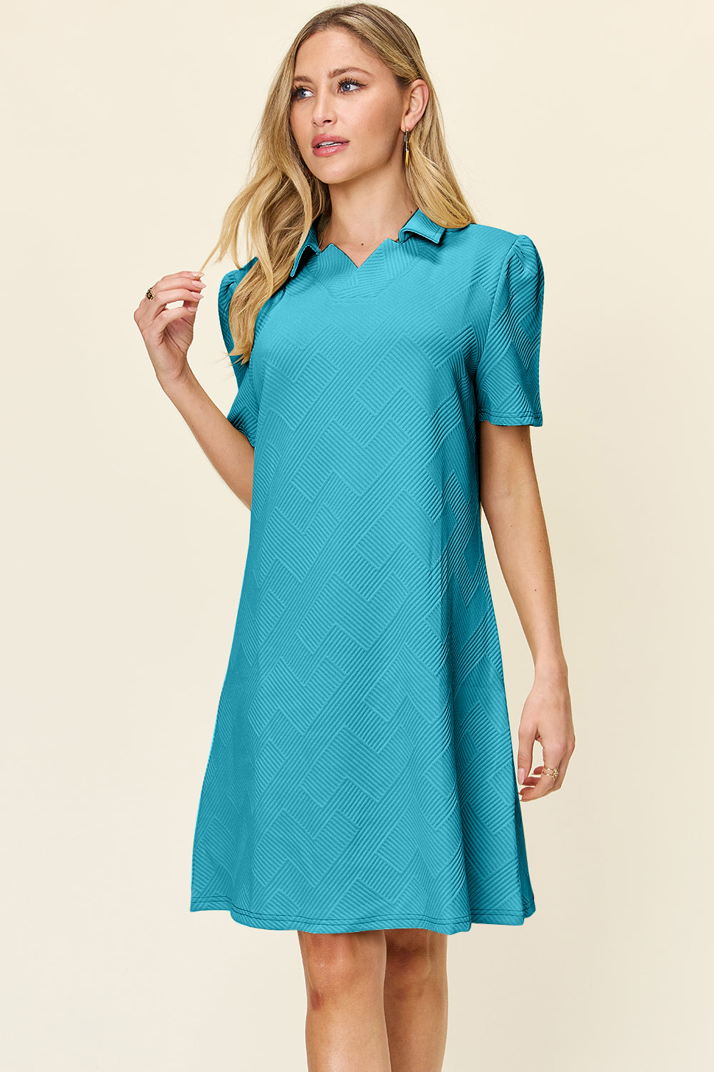 A woman poses against a plain background in the Double Take Full Size Texture Collared Neck Short Sleeve Dress, showcasing its geometric pattern and chic yet simple style. The dress, made from imported polyester, is textured in teal and features short sleeves. She has long blonde hair and gazes off to the side.