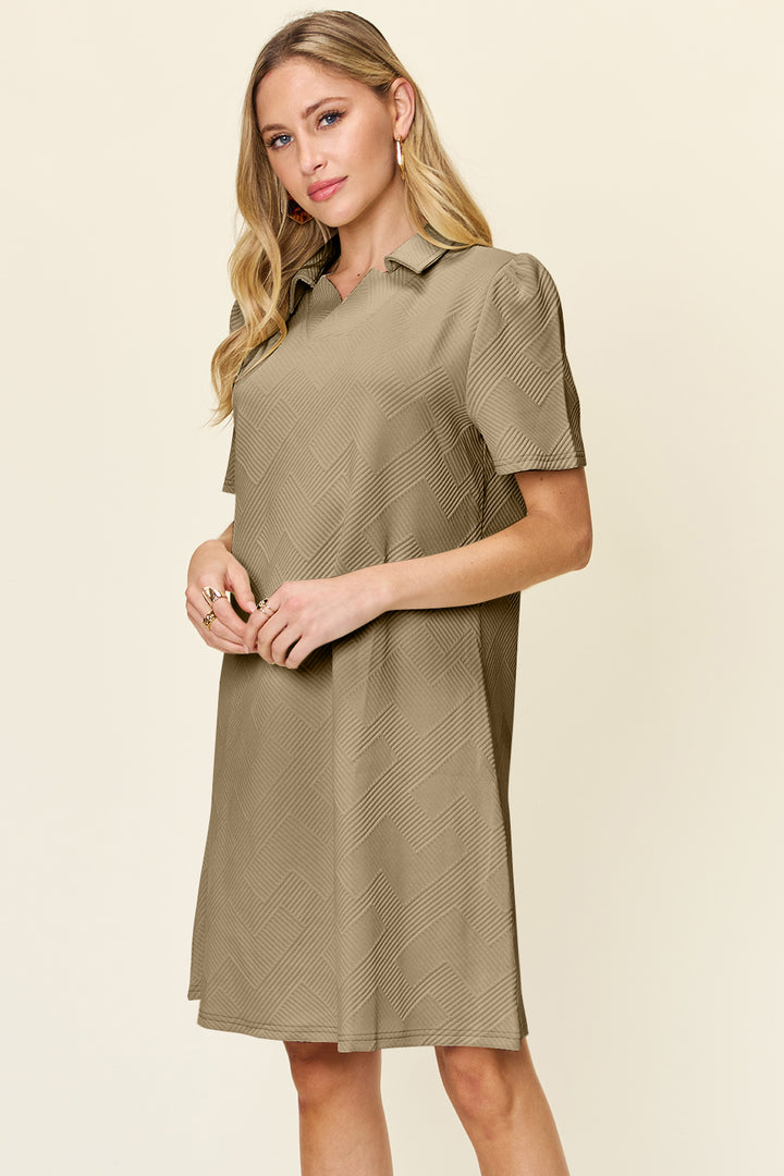 A woman with long blonde hair models the Double Take Full Size Texture Collared Neck Short Sleeve Dress, a knee-length, khaki piece crafted from imported polyester and adorned with a subtle zigzag pattern. She stands against a neutral background, slightly angled to the side, embodying a basic yet stylish look.