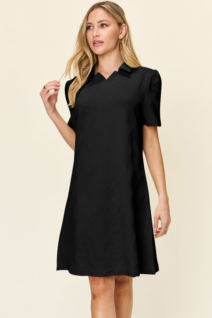 A woman with long blonde hair is wearing the Double Take Full Size Texture Collared Neck Short Sleeve Dress, designed in a basic style and crafted from imported polyester with a subtle textured pattern. She stands against a plain light-colored background, her left hand adjusting her hair, and a neutral expression gracing her face.