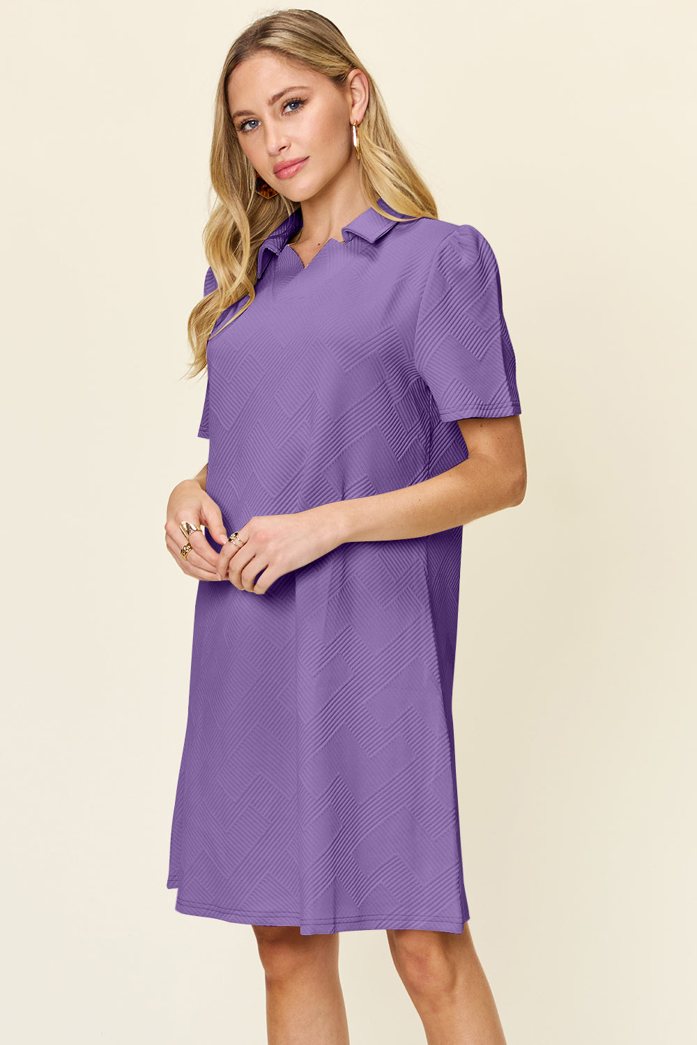 A person stands against a light background wearing the Double Take Full Size Texture Collared Neck Short Sleeve Dress. The dress, crafted from imported polyester and adorned with a subtle zigzag pattern, is short-sleeved and collared in a striking purple hue. The individual has long blond hair cascading down as they look slightly to the side, holding their hands together.