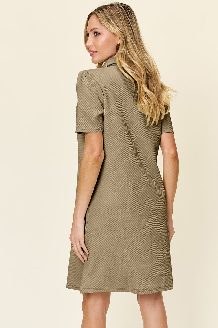 A woman with long blonde hair wears the Double Take Full Size Texture Collared Neck Short Sleeve Dress, which is knee-length and made of imported polyester in a basic style. The dress features a subtle zigzag pattern, and she stands facing away with her side profile visible against a plain light beige background.