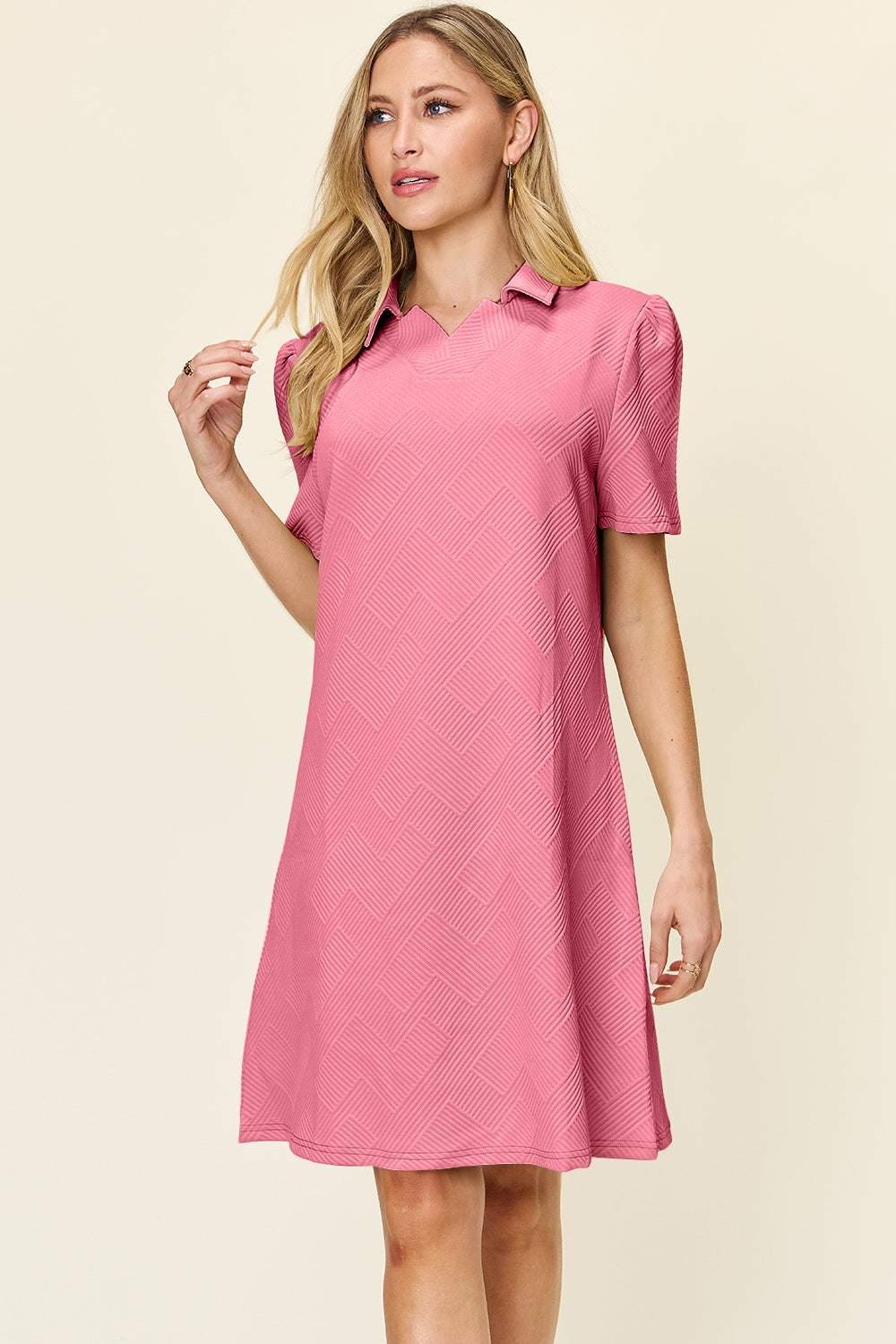 A woman wearing the Double Take Full Size Texture Collared Neck Short Sleeve Dress, crafted from imported polyester in a pink pattern with short puff sleeves, stands against a light background. Her long blonde hair cascades down as she looks to the side, gently touching her hair with one hand.