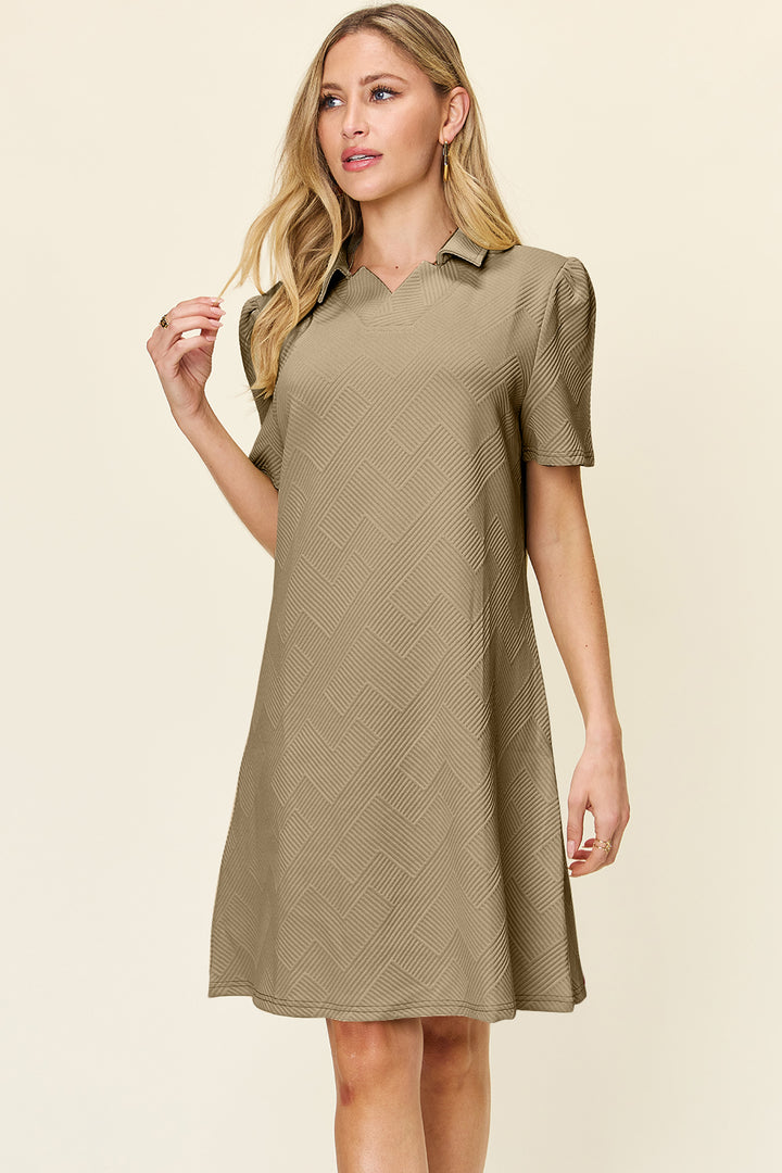 A woman with long, blonde hair is wearing the Double Take Full Size Texture Collared Neck Short Sleeve Dress, a knee-length, beige ensemble made of imported polyester. It features short puffed sleeves and a subtle geometric pattern. She stands against a plain light background, gazing slightly to the side while touching her hair in this stylish outfit.