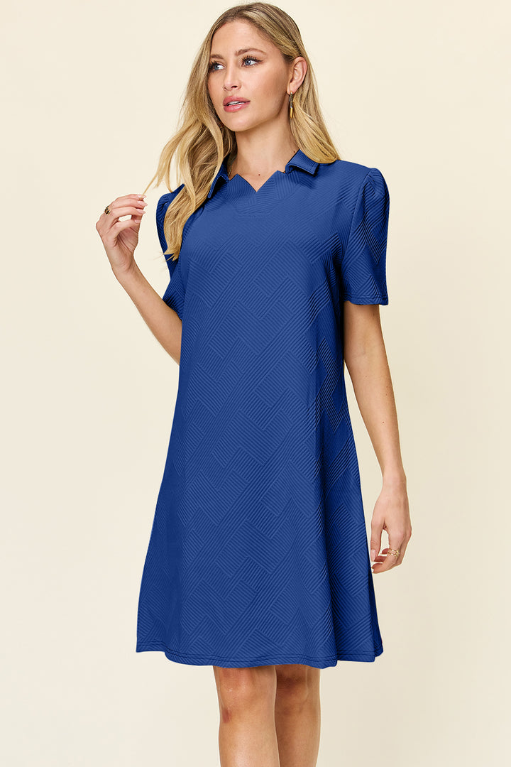 A woman with long blonde hair is wearing the imported "Double Take Full Size Texture Collared Neck Short Sleeve Dress," which features a basic style with a subtle geometric pattern. She stands against a plain background, looking slightly to the right.