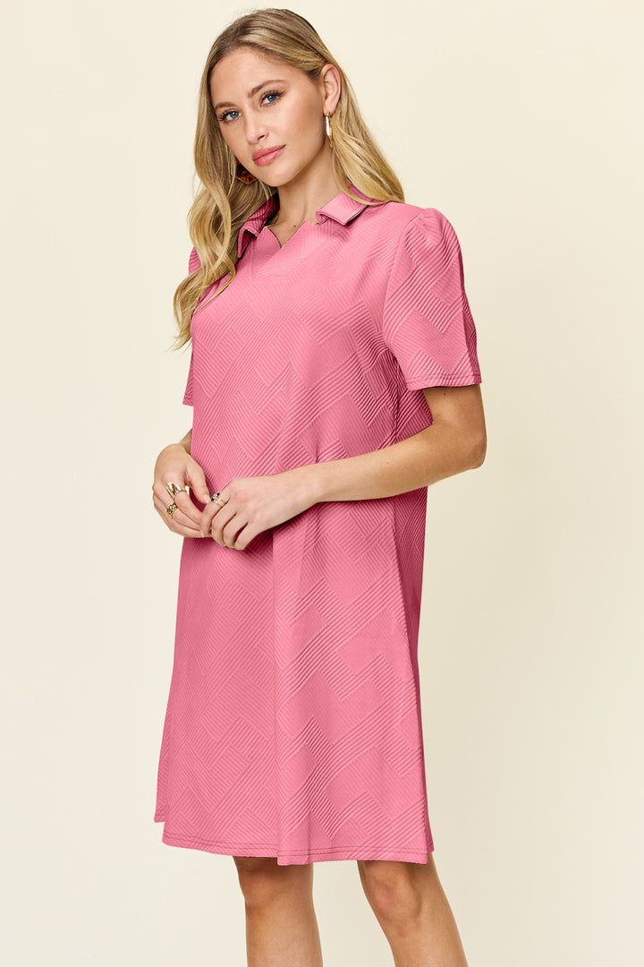 A woman in the Double Take Full Size Texture Collared Neck Short Sleeve Dress, featuring a subtle chevron pattern and made from imported polyester, stands against a plain background. Embracing basic style, she has long blond hair and poses with one hand near her waist, looking slightly to the side.