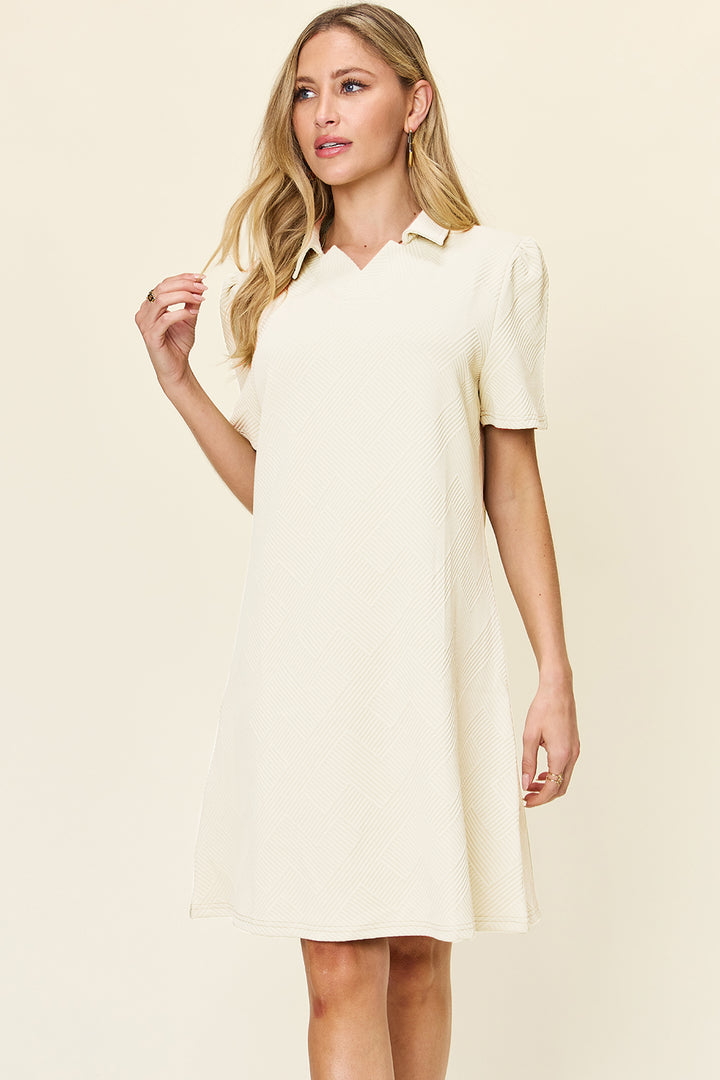 A woman with long blonde hair stands against a light background, wearing a Double Take Full Size Texture Collared Neck Short Sleeve Dress in cream. Made from imported polyester, she holds her hair with her right hand and gazes slightly to the side.