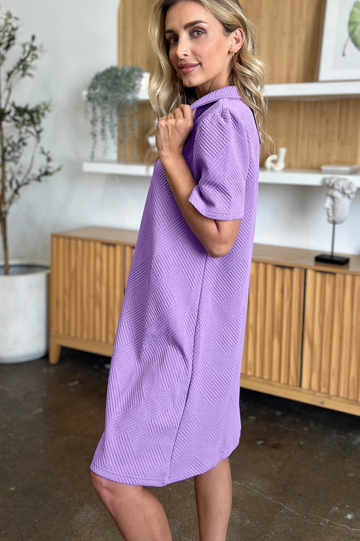 A person with long blonde hair is elegantly dressed in the "Double Take Full Size Texture Collared Neck Short Sleeve Dress," a knee-length purple polyester outfit. They stand in a room with minimalist decor, featuring a wooden cabinet and plants, smiling slightly as they glance over their shoulder.