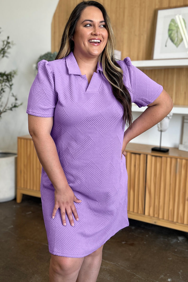 A person smiling and wearing the Double Take Full Size Texture Collared Neck Short Sleeve Dress in purple stands with one hand on their hip. The background features imported wooden furniture and a plant, creating an inviting scene.