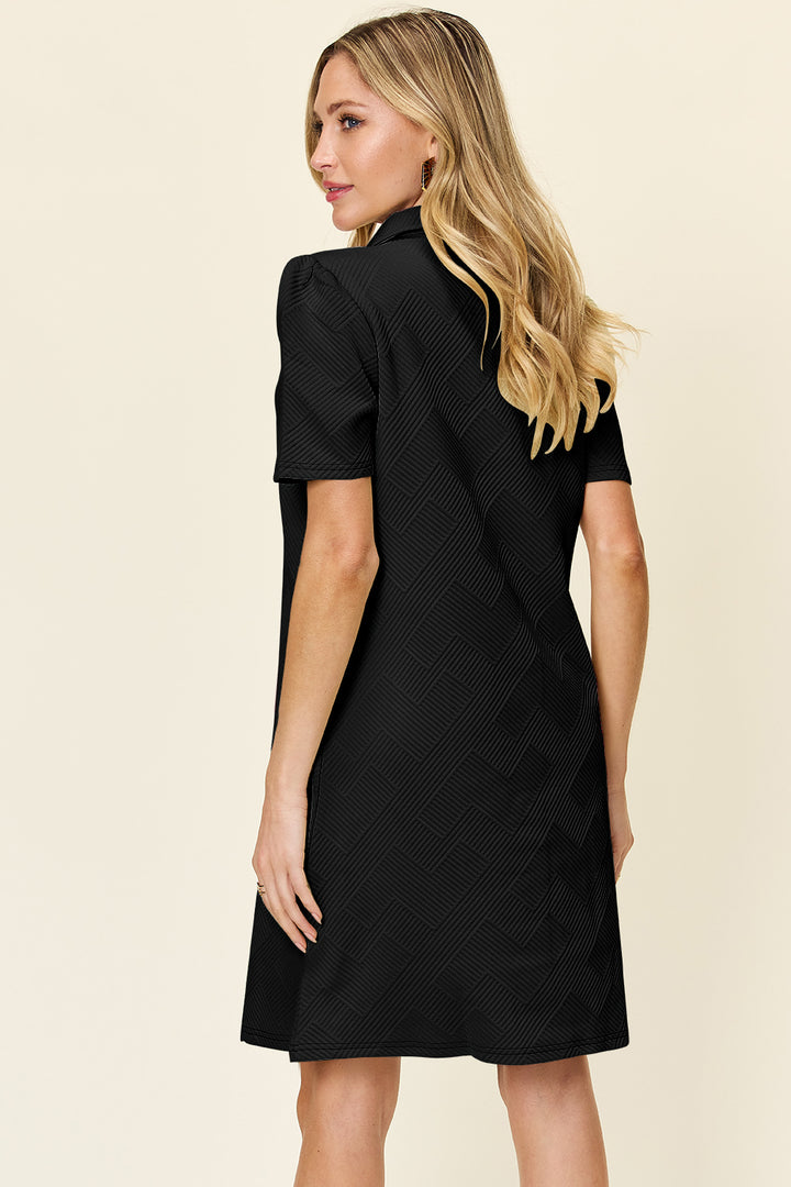 A woman with long, blonde hair showcases a basic style by wearing the "Double Take Full Size Texture Collared Neck Short Sleeve Dress" in a subtle zigzag pattern made from imported polyester. She is facing slightly away from the camera against a light beige background.