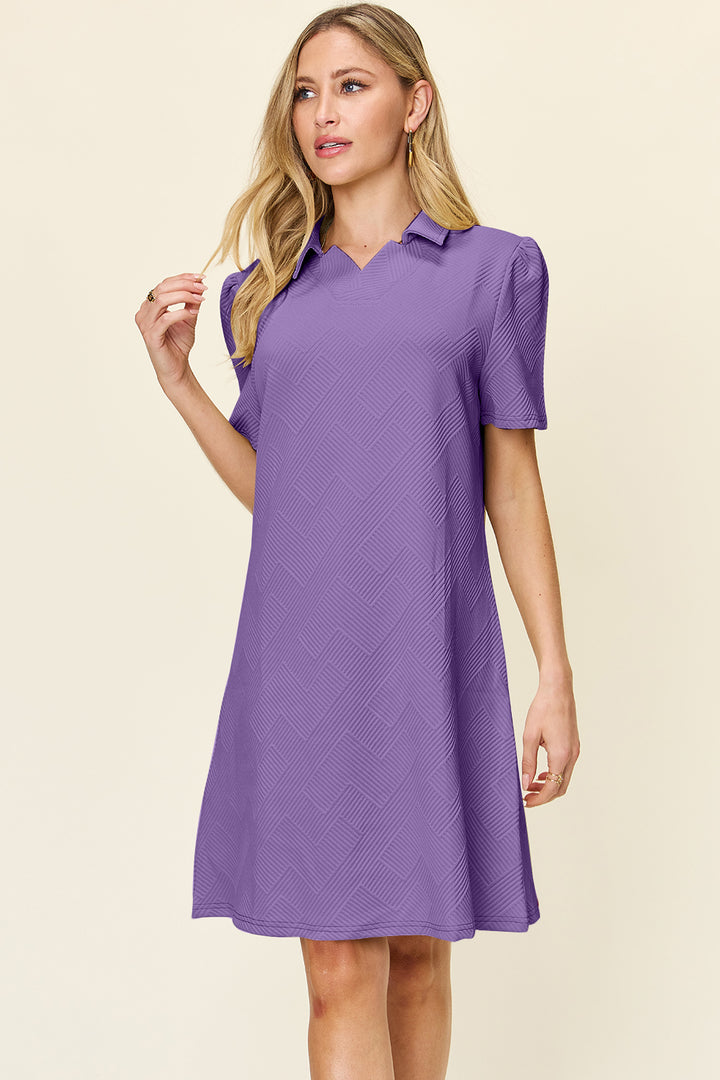 Wearing the Double Take Full Size Texture Collared Neck Short Sleeve Dress, a woman stands against a plain background. This knee-length dress in purple features a subtle geometric pattern that complements her long blonde hair, as she gazes to her right. Made from imported polyester fabric, the style exudes simplicity and elegance.