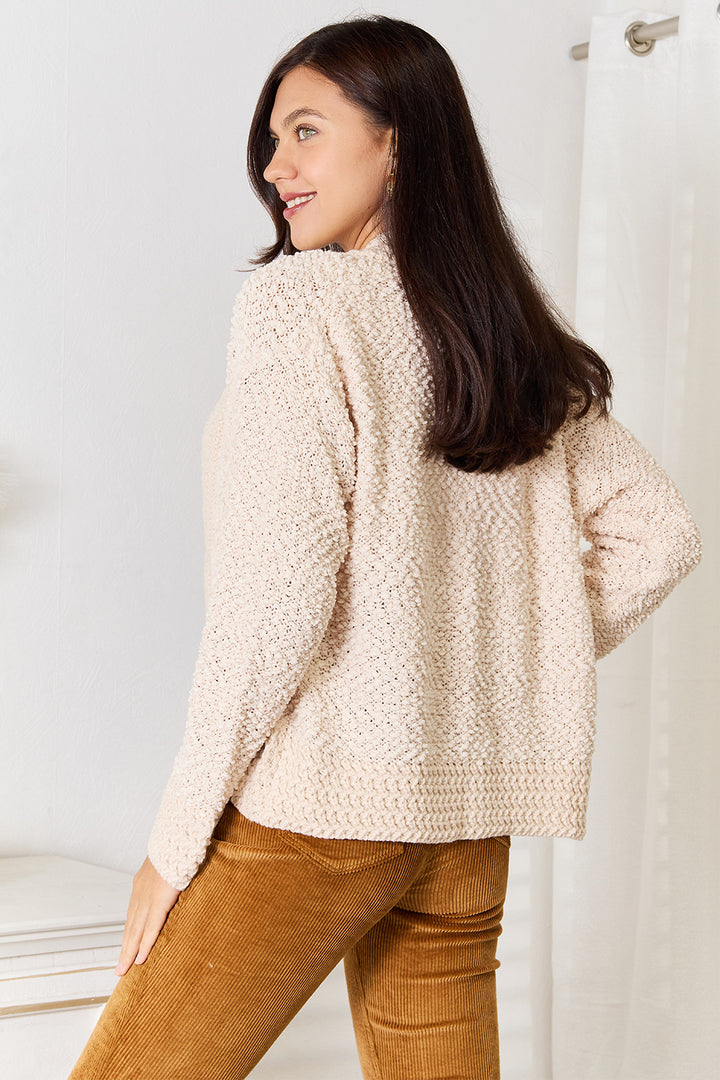 A woman with long dark hair is smiling and looking over her shoulder, wearing a cozy and versatile Double Take Open Front Long Sleeve Cardigan in beige, paired with mustard-colored corduroy pants. She stands near a white curtain and a light-colored surface, radiating effortless style.