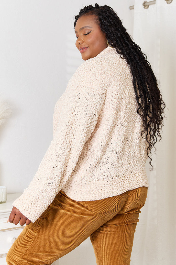 A woman with long curly hair is wearing the cozy and versatile Double Take Open Front Long Sleeve Cardigan layered over mustard-colored pants. She poses near a white wall and a curtain, her expression relaxed as she stands in the brightly lit room.