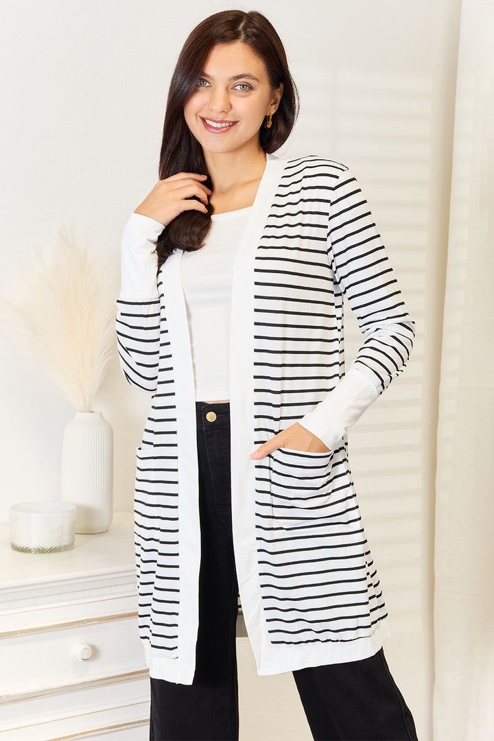 A woman smiles while wearing the Double Take Striped Open Front Longline Cardigan over a white top and black pants. She stands near a white dresser adorned with decorative items, with her hand casually tucked in one pocket.