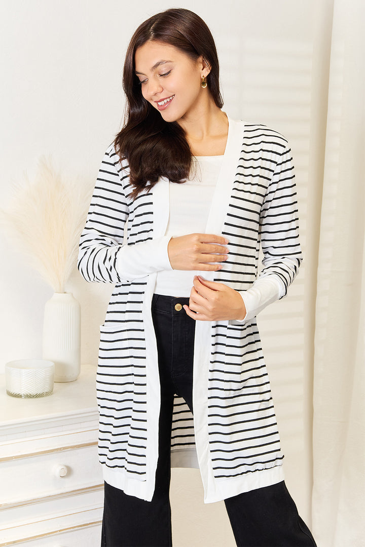 A woman wearing the Double Take Striped Open Front Longline Cardigan over a white top and black pants stands in a well-lit room, smiling with a relaxed pose. A small table with decorative items is visible in the background.