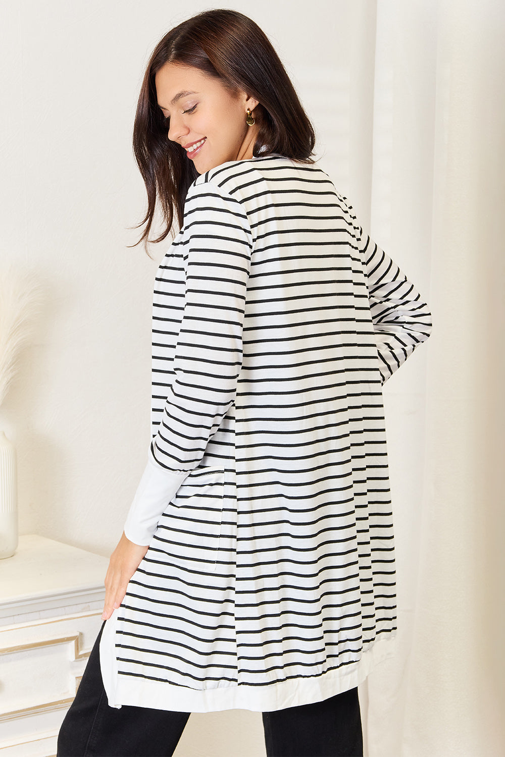 A woman with long dark hair, wearing the Double Take Striped Open Front Longline Cardigan, stands smiling in a bright room. She is looking over her shoulder, and a small table with decor is visible in the background.