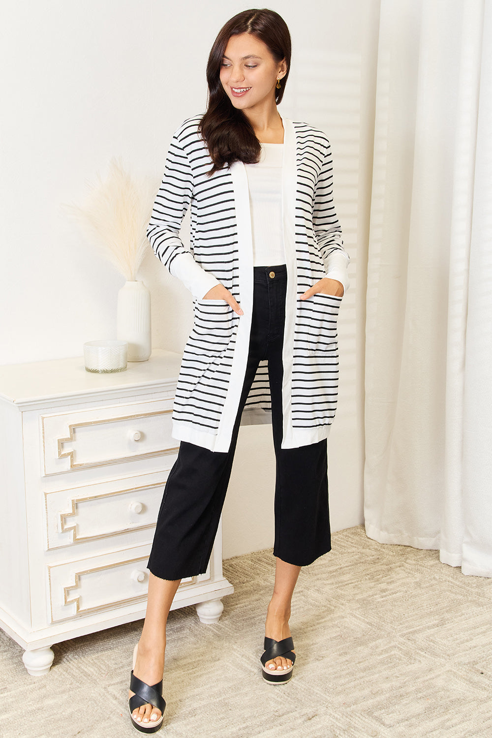 A woman stands in a room wearing a Double Take Striped Open Front Longline Cardigan over a white top, paired with black cropped pants and black sandals. She is smiling and standing near a white dresser adorned with decorative items.