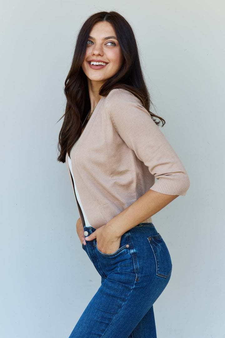 A smiling woman with long dark hair poses with one hand on her hip against a plain light background, wearing the Doublju My Favorite Full Size 3/4 Sleeve Cropped Cardigan in Khaki paired with blue jeans.