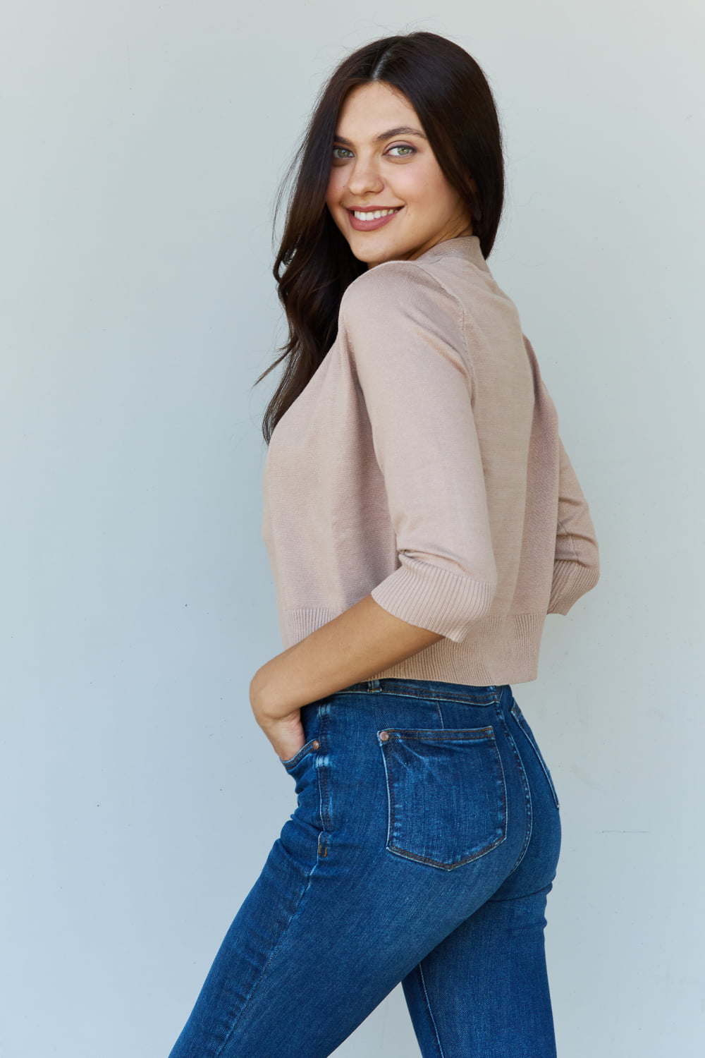 A woman with long dark hair smiles while looking over her shoulder, wearing the Doublju My Favorite Full Size 3/4 Sleeve Cropped Cardigan in Khaki and blue jeans against a plain light background.