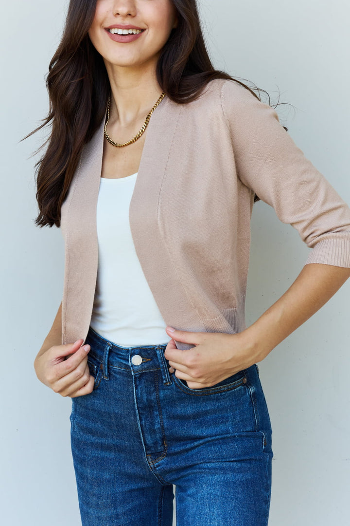 A person with long, dark hair is wearing the Doublju My Favorite Full Size 3/4 Sleeve Cropped Cardigan in Khaki over a white top and blue jeans. They are smiling at the camera while adjusting the stylish cardigan with one hand. The plain, light-colored background highlights their relaxed yet chic look.