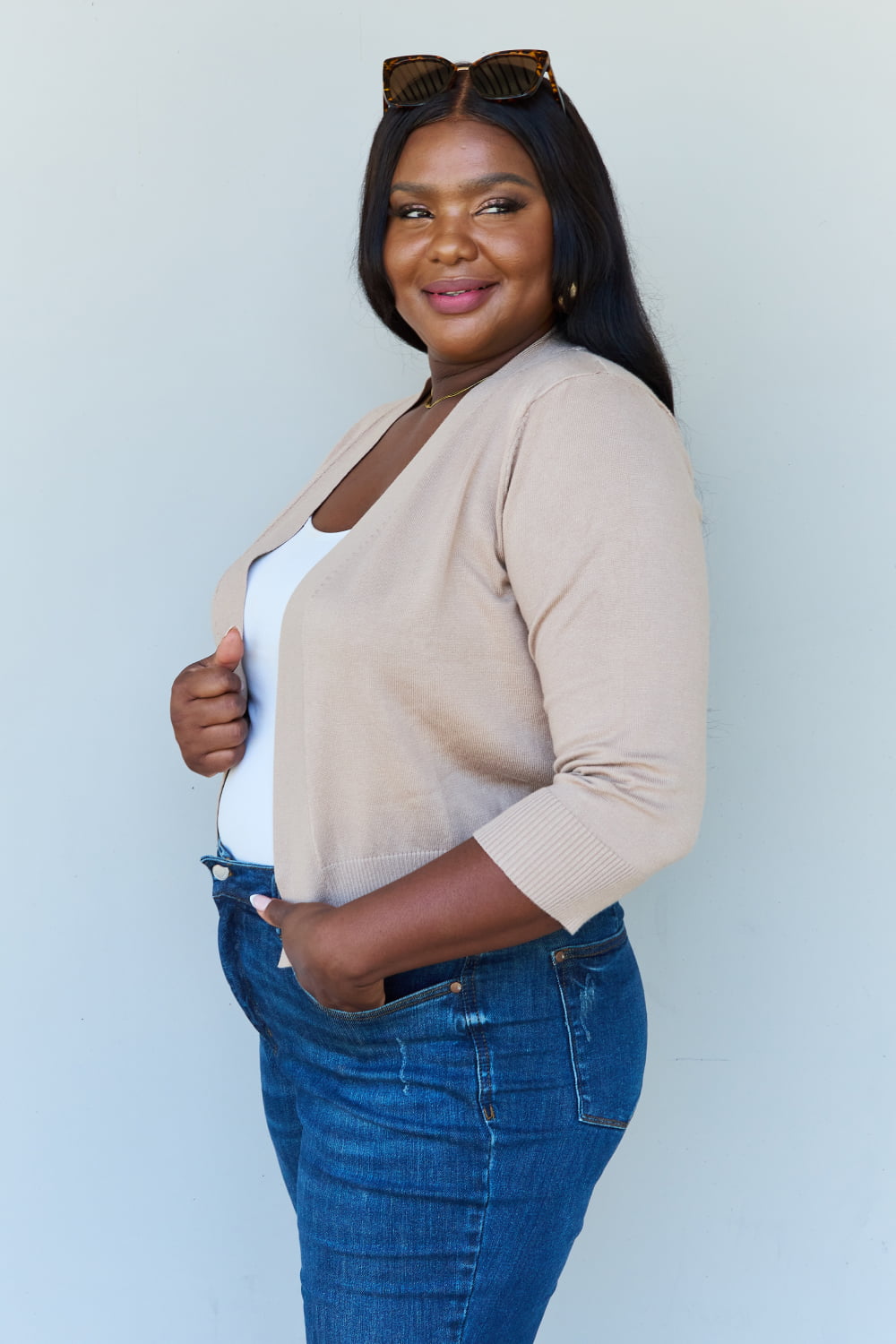 A woman stands smiling against a neutral background, wearing sunglasses and the Doublju My Favorite Full Size 3/4 Sleeve Cropped Cardigan in Khaki over her white shirt and blue jeans. Her hands rest near her pockets, and her long dark hair cascades loosely over her shoulders.