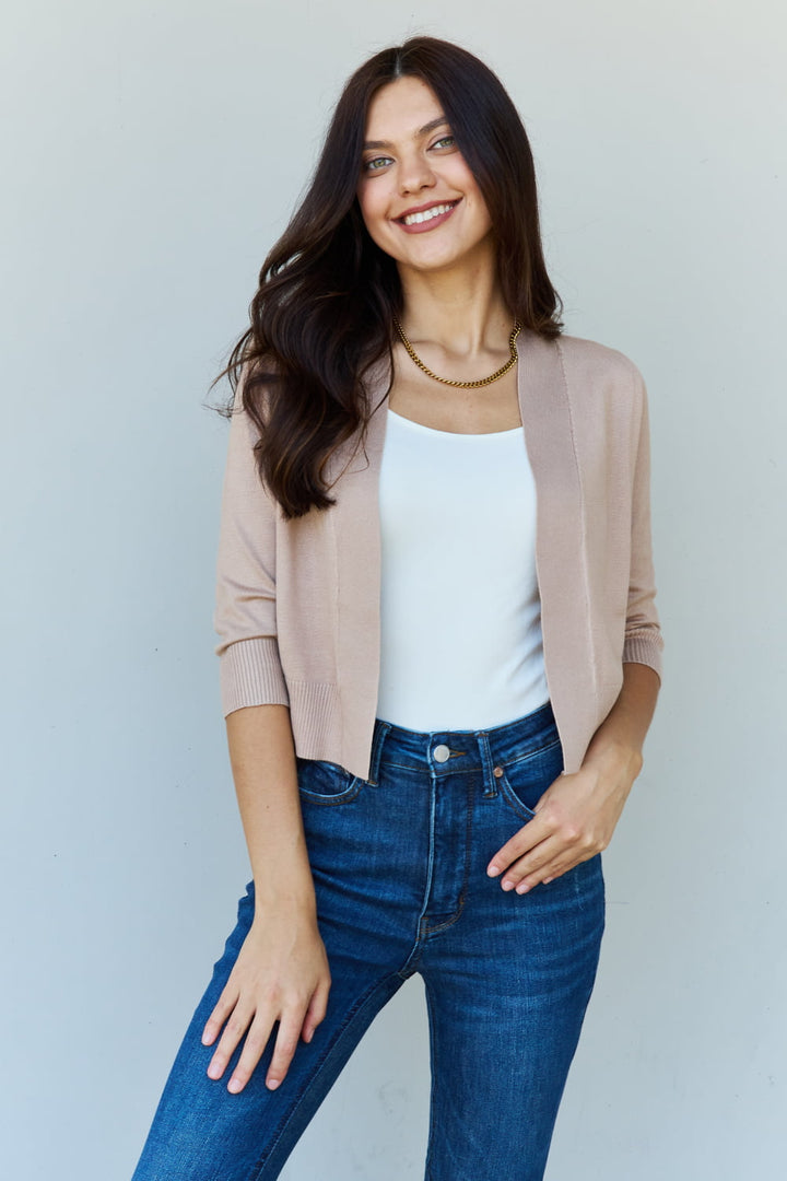 A woman smiles while standing against a plain background, wearing the Doublju My Favorite Full Size 3/4 Sleeve Cropped Cardigan in khaki over a white top and blue jeans.