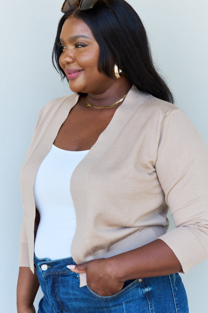 A person with long dark hair and gold hoop earrings is wearing sunglasses on their head, the Doublju My Favorite Full Size 3/4 Sleeve Cropped Cardigan in Khaki, a white top, and blue jeans. They are smiling and looking to the side.