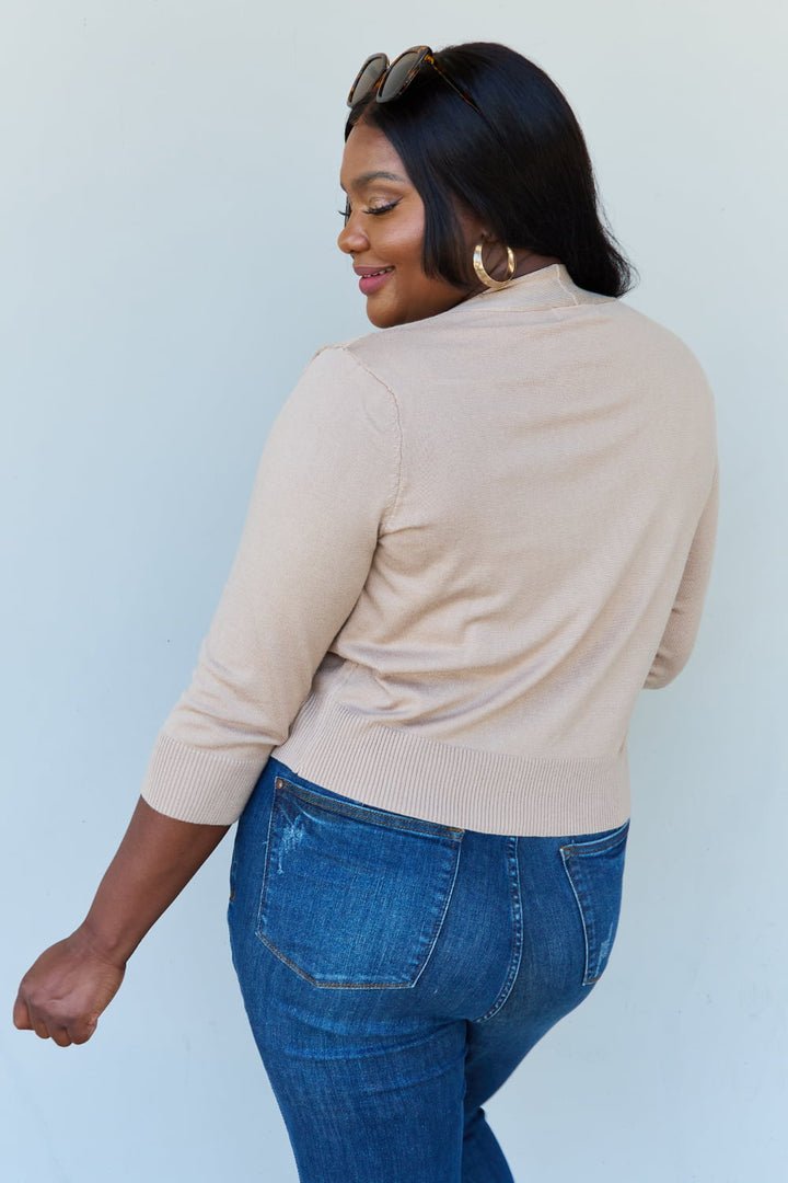 A woman with long dark hair is slightly turned away, wearing the Doublju My Favorite Full Size 3/4 Sleeve Cropped Cardigan in Khaki along with blue jeans. She has sunglasses on her head and is smiling against a plain light background.