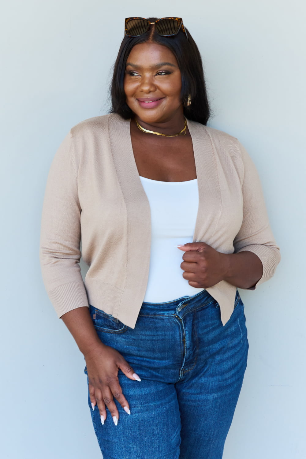 In front of a simple backdrop, a woman exudes confidence with her warm smile and gently clasped hands. She's wearing the Doublju My Favorite Full Size 3/4 Sleeve Cropped Cardigan in Khaki over a white top and paired with blue jeans. Her look is further complemented by sunglasses perched on her head and gold jewelry accents.