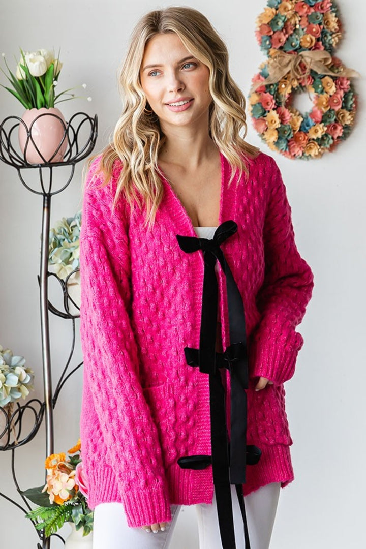 A person wearing the First Love Tie Closure Open Knit Cardigan in a bright pink, relaxed fit with black ribbon details stands indoors. In the background, a decorative floral wreath graces the wall alongside flower arrangements in pastel vases.