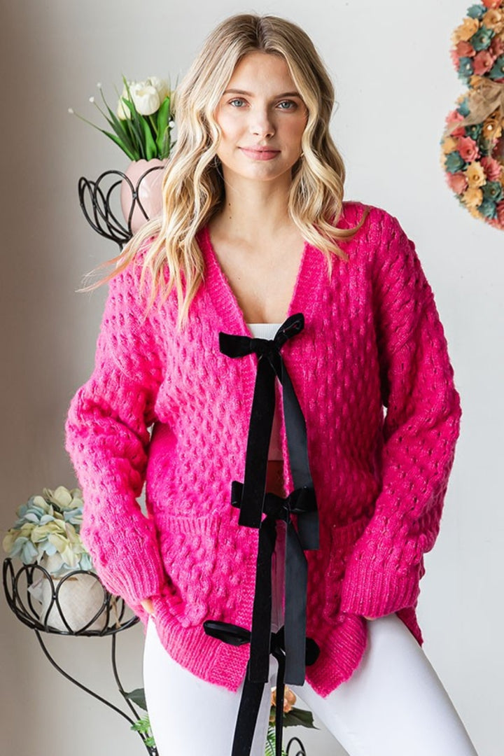 A person with long blonde hair is wearing the First Love Tie Closure Open Knit Cardigan in bright pink, featuring comfortable black ribbon ties. They're smiling with hands tucked into their pockets, standing in a room decorated with flowers and a plant.