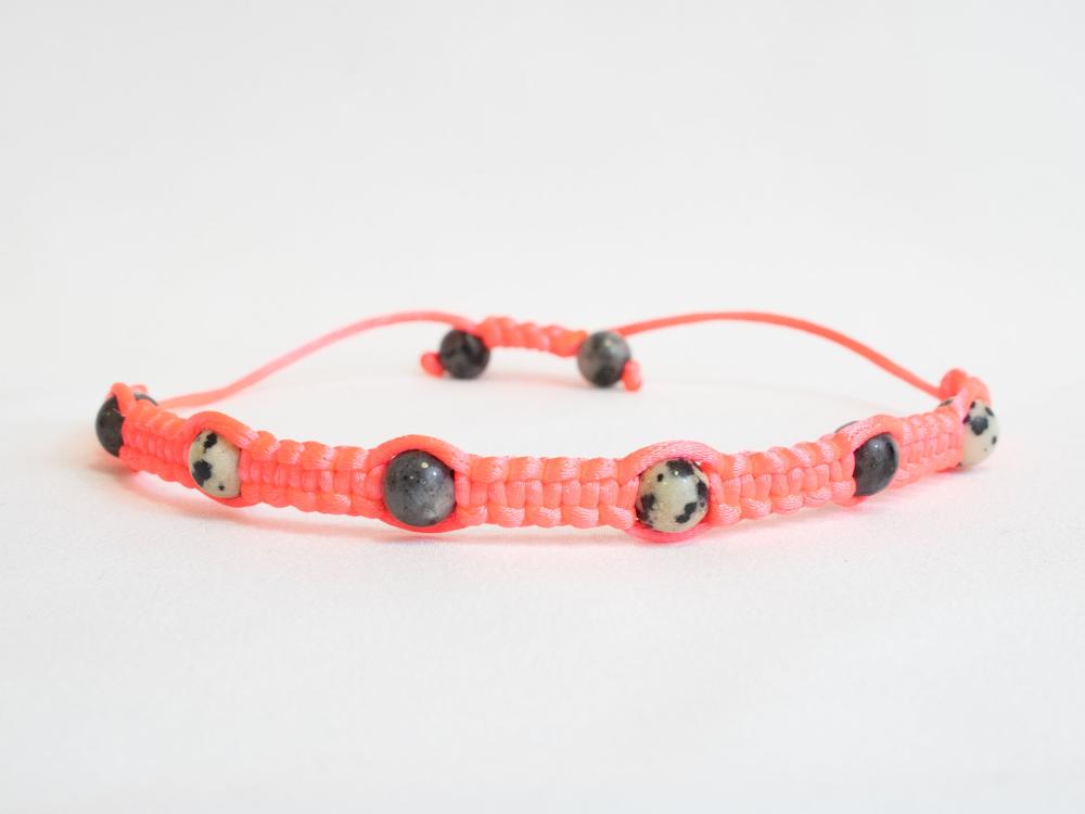 The Gem and Stone Beach Bracelet by Claire is a unique piece, showcasing black and white speckled beads artfully woven with vibrant pink thread. This handcrafted bracelet offers versatility with its adjustable closure, presented elegantly against a plain, light backdrop.