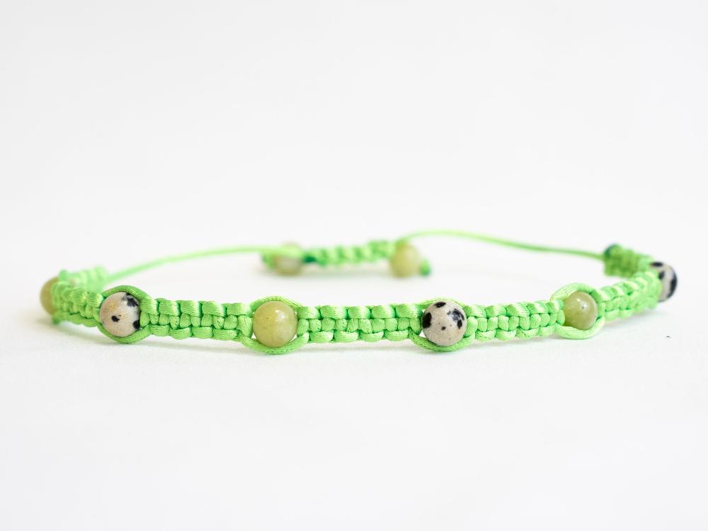 Introducing the Gem and Stone Beach Bracelets by Claire: A unique bracelet that boasts a green macramé cord adorned with round, spotted beads. Its adjustable closure ensures a perfect fit for any wrist. Set against a plain white background, the vibrant colors of this standout accessory truly shine.