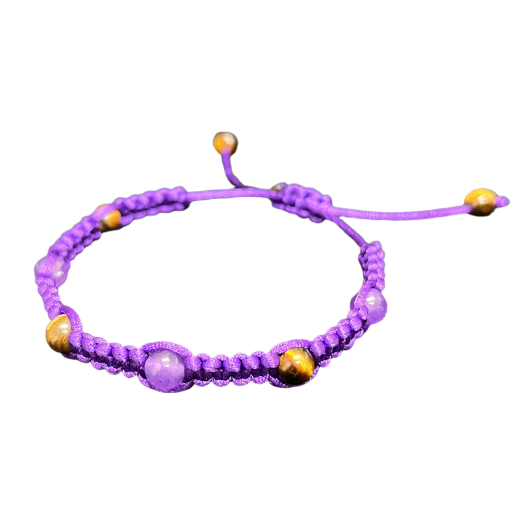 The unique Gem and Stone Beach Bracelet by Claire, featuring a purple braided design interspersed with luxurious purple and gold beads, is elegantly displayed on a dark fabric background.